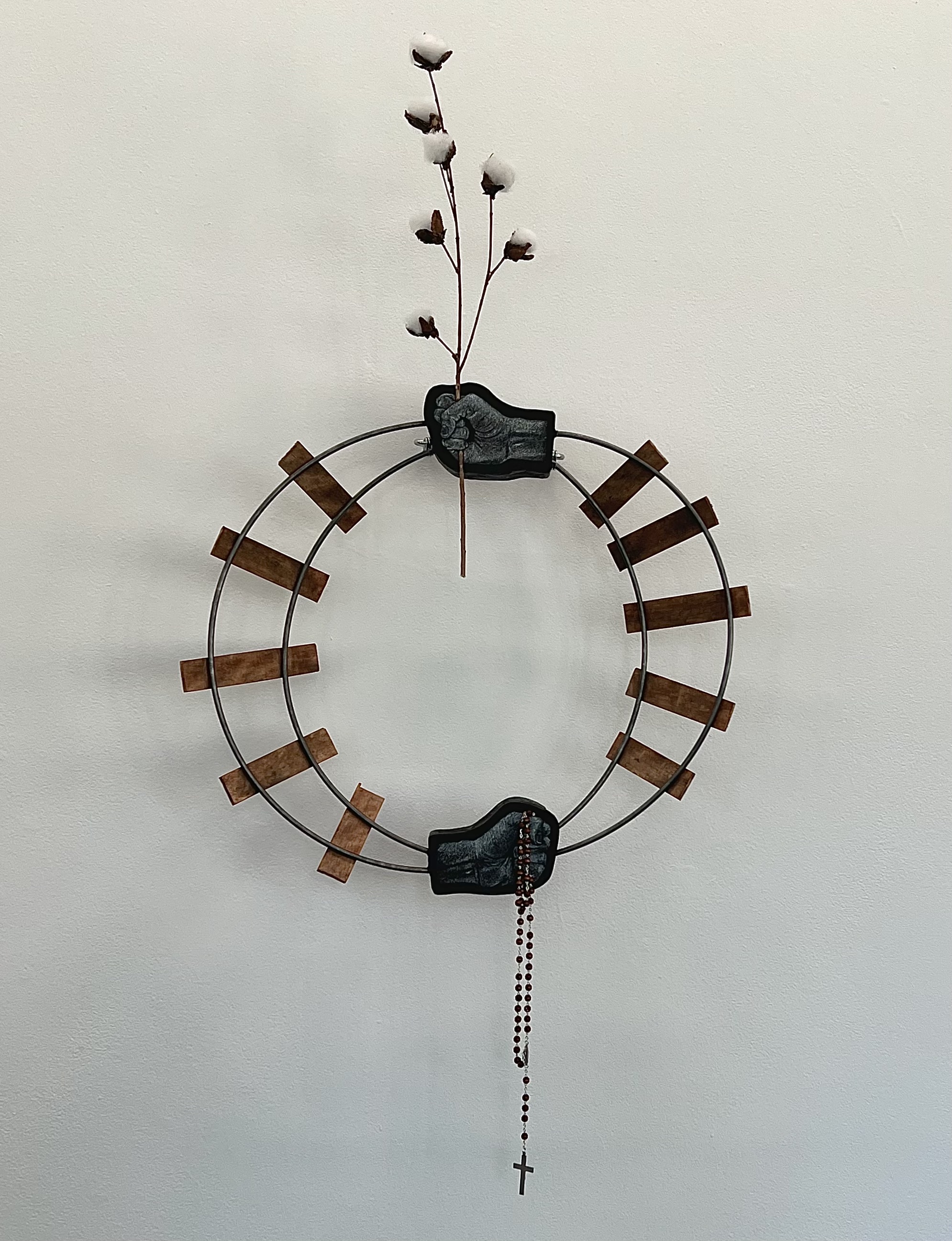 Metal, wood, and plant sculpture in the shape of a circle, with hands grasping objects near the top and bottom